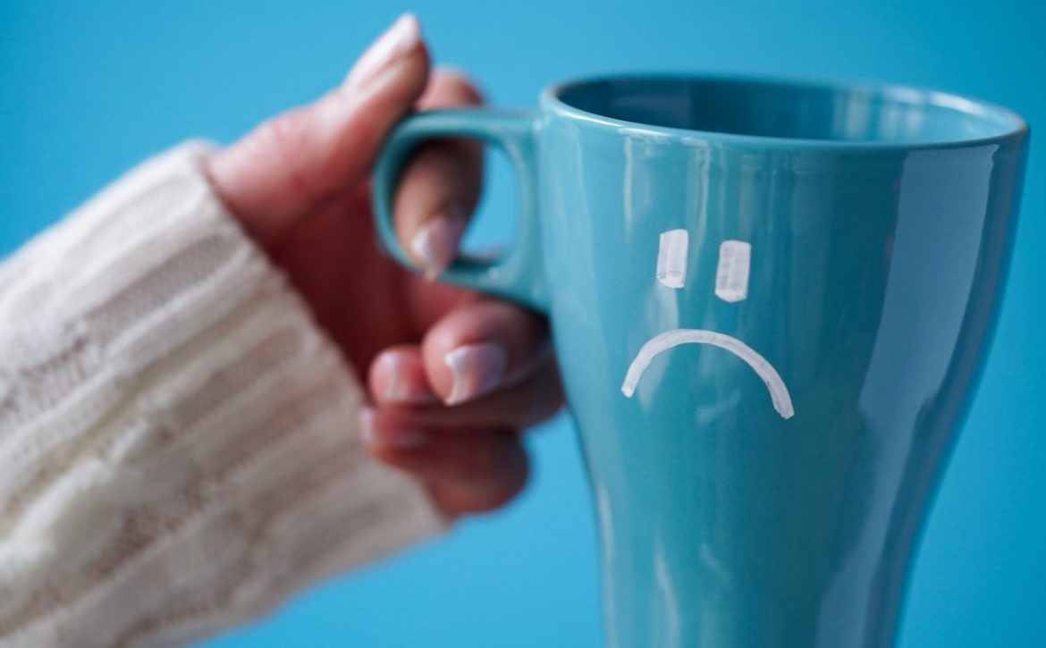 Blue Monday concept. Close-up of a female hand in a white sweater holds a blue cup with a sad smiley as a symbol of the most depressive day of the year on a blue background.