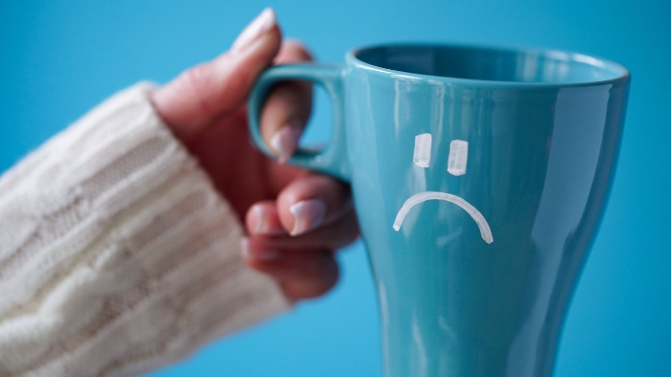 Blue Monday concept. Close-up of a female hand in a white sweater holds a blue cup with a sad smiley as a symbol of the most depressive day of the year on a blue background.