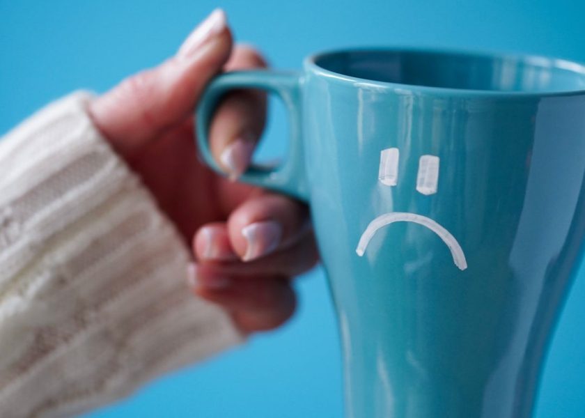 Blue Monday concept. Close-up of a female hand in a white sweater holds a blue cup with a sad smiley as a symbol of the most depressive day of the year on a blue background.