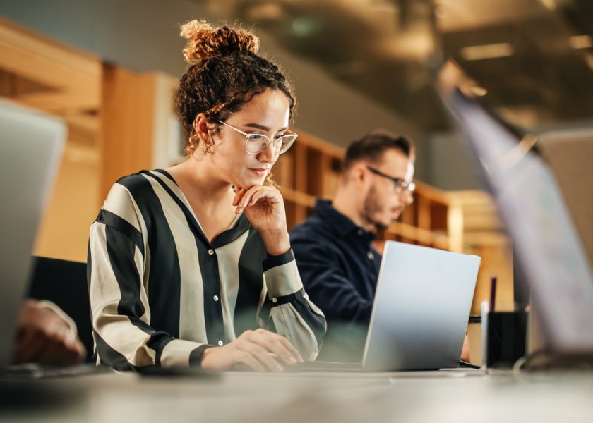 Portrait of Enthusiastic Hispanic Young Woman Working on Computer in a Modern Bright Office. Confident Human Resources Agent Smiling Happily While Collaborating Online with Colleagues.